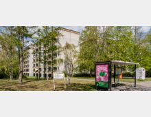 Mehrstöckiges Wohngebäude mit Balkonen in einer grünen, parkähnlichen Umgebung; im Vordergrund eine überdachte Bushaltestelle mit einer Werbetafel sowie Bäume und gepflegte Rasenflächen.