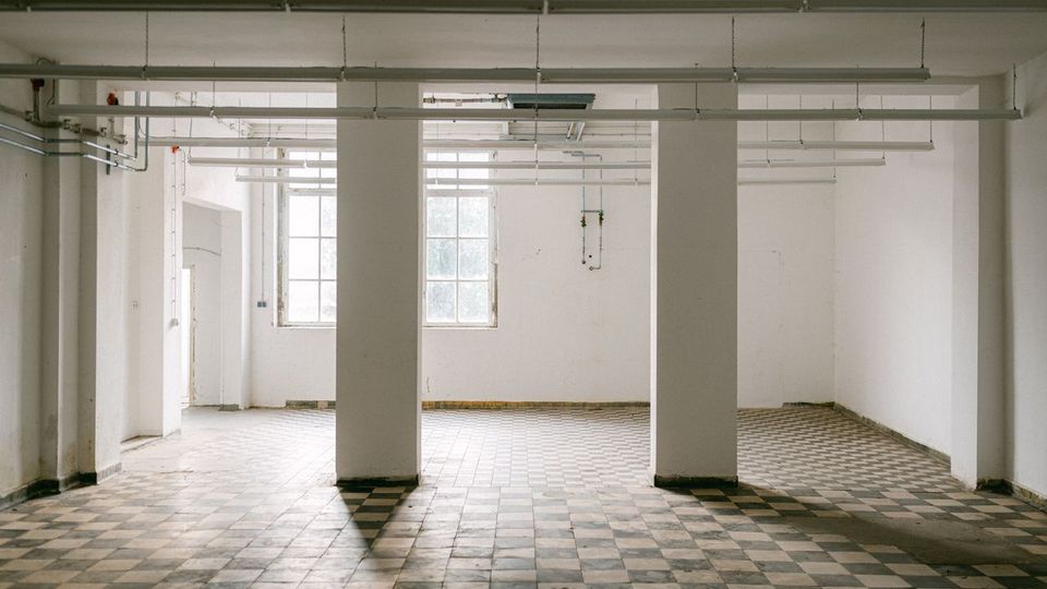 Leerer, heller Industrie- oder Lagerraum mit weiß gestrichenen Wänden, vier rechteckigen Stützpfeilern und einem schwarz-weiß karierten Fliesenboden. An der Decke verlaufen lange Leuchtstoffröhren und Leitungen; hinten sind große Fenster, die Tageslicht hereinlassen.