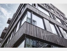 Moderne Gebäudefassade mit dunklem Holz verkleidet, großen Glasfenstern und Balkonen. Der Himmel ist teilweise bewölkt, und in den Fensterscheiben spiegeln sich Umgebung und bauliche Details.