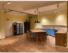 Eine moderne Gemeinschaftsküche mit gelben Wänden, Holzfußboden und einer zentralen Kücheninsel mit Barhockern. An der Wand befinden sich blaue Küchenschränke, ein Kühlschrank, und offene Regale, die mit minimalistischer Hängeleuchte beleuchtet werden.