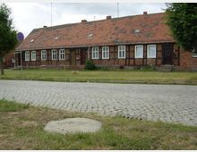 Fachwerk-Reihenhaus mit rotem Ziegeldach, kleinen Dachfenstern und weißen Sprossenfenstern in ländlicher Umgebung. Im Vordergrund eine gepflasterte Straße und eine Rasenfläche.