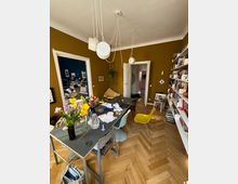 Esszimmer mit senfgelben Wänden, einem Holzboden in Fischgrätmuster und einem großen Tisch, der mit Papieren, Blumen und Gegenständen bedeckt ist. Im Raum befinden sich außerdem moderne Stühle, eine Bücherwand und eine Hängeleuchte mit mehreren Kabeln und Lampen. Türen führen in angrenzende Räume, darunter ein Bereich mit dunkelblauer Wandfarbe.