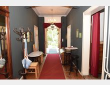 Ein länglicher Flur mit dunklen Wänden und Holzfußboden, dekoriert mit einem roten Teppich. An den Wänden befinden sich kleine Bilder, links stehen ein Garderobenständer und eine kleine Sitzgruppe mit einem Marmortisch, am Ende des Flurs hängt ein Kronleuchter und die doppelflügelige Tür öffnet sich zu einem Garten.