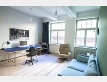 Helles Büro mit Holzboden, einem großen Schreibtisch mit Computer und Lampe, einem schwarzen Bürostuhl und einer beige Ledersessel. Im Hintergrund befinden sich zwei große Fenster, ein blaues Sofa und dunkle Vorhänge.