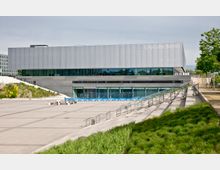 Modernes Veranstaltungsgebäude mit der Aufschrift "CityCube Berlin", rechteckige Architektur mit einer metallischen oberen Fassade und großen Glasfenstern im unteren Bereich. Vor dem Gebäude liegt ein gepflasterter Platz mit Sitzbänken, umgeben von Grünflächen und Treppen.