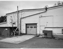 Gebäuderückseite mit glatter Betonfassade, einem Rolltor in der Mitte und freiliegenden Rohren sowie Verkabelungen. Links befindet sich ein kleiner Anbau mit Wellblechverkleidung, im Vordergrund Asphaltfläche. Ein kahler Baum und vereinzelte Blätter ergänzen die Szene.