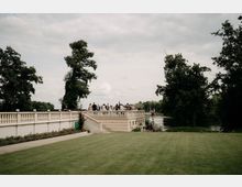Weiße steinerne Terrasse mit Balustrade und Treppe in einem Park, davor eine große Rasenfläche. Rechts verläuft ein Fluss entlang dichter Bäume, im Hintergrund sind weitere Grünflächen unter bewölktem Himmel zu sehen.