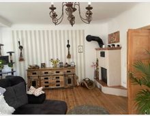 Wohnzimmer mit hellem Holzboden, gemusterter Tapete und einem weißen Kamin mit schwarzem Ofenrohr. Im Raum stehen eine rustikale Holzkommode, ein schwarzes Sofa, eine Katzenspielstruktur sowie Deko-Objekte wie Pflanzen und Bilder.