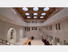 Großes, modernes Atrium mit hellen Wänden, umlaufenden Galerien und vielen rechteckigen Fenstern auf mehreren Ebenen. Die Decke ist mit runden Oberlichtern und einer perforierten Holzverkleidung gestaltet; im Erdgeschoss stehen Bänke und Sitznischen neben hohen Rundbögen und Glasfronten.