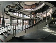 Modernes Atrium mit einer großen, geschwungenen Treppe aus Stahl und Glasgeländern in einem offenen, runden Raum. Umgeben von mehreren Stockwerken mit großen Glasfenstern, die viel Tageslicht hereinlassen.