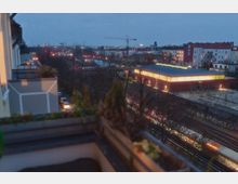 Blick von einem Balkon auf eine städtische Umgebung in der Dämmerung mit beleuchteten Gebäuden, eisenbahngleisen und Straßen. Im Vordergrund ein Pflanzkasten, während ein modernes rotes Gebäude mit großen Fenstern im rechten Bildbereich hervortritt.