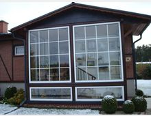 Außenansicht eines Hauses mit einem großen Wintergarten, dessen Fassade aus großen, weißen Sprossenfenstern besteht. Das Gebäude hat eine Backsteinfassade, ein dunkles Satteldach und ist von einem kleinen, verschneiten Garten mit dekorativen Büschen umgeben.