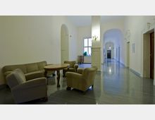 Heller Flur in einem historischen Gebäude mit glänzendem Fliesenboden, gewölbten Türrahmen und einer Sitzecke mit vier gepolsterten Sesseln, einem Sofa und einem runden Holztisch. Im Hintergrund sind ein Fenster, eine Wandleuchte und eine geöffnete Verbindungstür sichtbar.