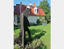 Skulptur aus Metall in einem gepflegten Garten mit grünem Rasen, vor einem weißen Haus mit roten Dachschindeln und zwei Gaubenfenstern. Im Hintergrund sind Büsche und Bäume sowie Gartenmöbel sichtbar.