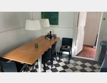 Ein kleiner Essbereich mit einem rechteckigen Holztisch, umgeben von schwarzen Stühlen, steht auf einem schwarz-weiß karierten Fliesenboden. Zwei weiße Lampenschirme hängen über dem Tisch, und im Hintergrund befindet sich eine geöffnete Holztür, die zu einem Raum mit roten Fliesen führt.