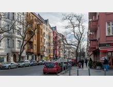 Straßenszene in einer Stadt mit mehrstöckigen Wohngebäuden in verschiedenen Farben, darunter weiße, gelbe und rote Fassaden. Am Straßenrand parken Autos, während Fußgänger auf dem Gehweg entlanglaufen; vereinzelte Bäume ohne Laub sind zu sehen.