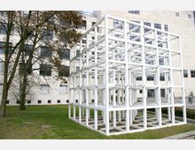Kubusartige, weiße Metallskulptur in einem modernen, begrünten Außenbereich vor einem großen, sandfarbenen Gebäude mit vielen Fenstern. Links steht ein kahler Baum, und im Hintergrund sind Gebäudefassaden sichtbar.