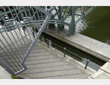 Treppenaufgang aus Metall mit Geländer neben einer schmalen Wasserstraße, die von einer Brückenkonstruktion mit grünen Stahlträgern überquert wird. Der Boden besteht aus gepflastertem Stein, abgegrenzt durch Metallpoller und Geländer.
