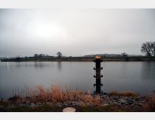 Flussufer mit ruhigem Wasser und einem metallischen Poller in der Mitte des Flusses. Im Vordergrund sind Gras und Felsen zu sehen, während im Hintergrund eine grasbewachsene Böschung mit vereinzelten Bäumen sichtbar ist.