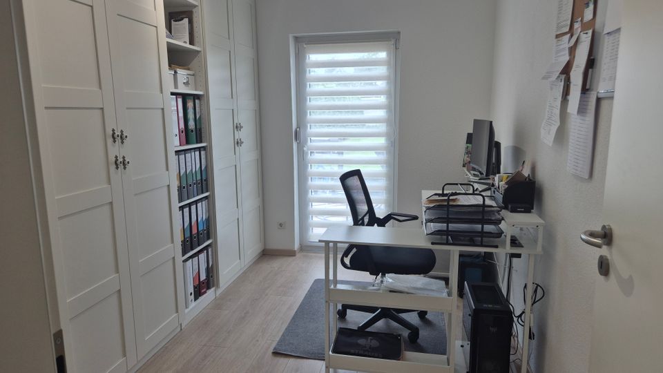 Schmales, helles Homeoffice mit hellem Holzfußboden, großem weißen Einbauschrank und Regalen mit Ordnern an der linken Wand. Rechts steht ein weißer Schreibtisch mit Computer und Ablagen vor einem Fenster mit Doppelrollo; an der Wand hängt ein Pinnbrett mit Zetteln.