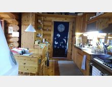 Küche in einem Holzhaus mit Wänden aus massiven Holzstämmen, einem Esstisch aus hellem Holz, hölzernen Schränken und Arbeitsflächen. Im Hintergrund hängt ein blauer Vorhang mit Hasenmotiven, davor befinden sich Küchenutensilien und Dekorationen.