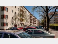 Straßenszene mit mehrstöckigen Wohnhäusern im Hintergrund, die eine helle Fassade und rote Zierelemente haben. Im Vordergrund stehen zahlreiche geparkte Autos entlang der Straße, und Bäume mit frischem Frühlingslaub säumen den Gehweg.
