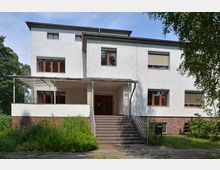 Wohngebäude im modernen Stil mit hellem Putz und roten Fensterrahmen. Das zweigeschossige Gebäude hat eine überdachte Eingangstreppe und ist von einer Grünfläche umgeben.