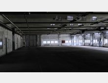 Eine leere industrielle Lagerhalle mit sichtbaren Stahlträgern an der Decke, mehreren Fenstern an den Seitenwänden und einer großen zweiflügeligen Tür an der Stirnseite. Die Halle wirkt abgedunkelt, mit spärlichem Tageslicht, das durch die Fenster einfällt.