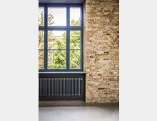 Innenraum mit großer, dunkel gerahmter Fensterfront und Blick auf grüne Bäume; darunter ein breiter Heizkörper. Rechts eine freiliegende Ziegelwand und ein glatter Betonboden im Vordergrund.