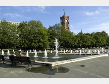Ein öffentlicher Platz mit Springbrunnen und Sitzbänken, umgeben von Bäumen und einem gepflasterten Boden, im Hintergrund ein historischer roter Turm mit Uhr und Fahne. Menschen sitzen auf den Bänken und entspannen im grünen Park.