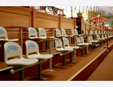 Reihen von weiß-blauen Kunststoffsitzen mit kleinen Tischen in einer Bowlingbahn-Lounge, umgeben von Holzvertäfelungen und dekorativen Elementen wie Girlanden. Im Hintergrund sind Bowlingkugeln und ein Bereich mit Stehtischen zu sehen.