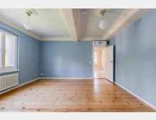 Leerer Wohnraum mit hellblauen Wänden und Holzdielenboden, links ein großes Fenster mit Heizkörper darunter. Die Decke hat dekorative Stuckelemente und zwei runde Hängelampen; rechts führt eine offene Tür in einen angrenzenden Flur.