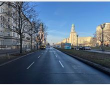 Breite, mehrspurige Stadtstraße mit Mittelstreifen, gesäumt von mehrstöckigen Altbauten und kahlen Straßenbäumen. Im Hintergrund ist ein markantes, hohes Gebäude mit Turm und grüner Kuppel zu sehen, daneben weitere große Verwaltungsgebäude unter blauem Himmel.