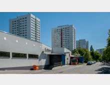 Industriegebiet mit einem modernen, grauen Lagergebäude im Vordergrund, daneben ein orangefarbener Lkw. Im Hintergrund stehen mehrere hohe Wohnhochhäuser mit Balkonen, umgeben von Bäumen und einer klaren, sonnigen Straße.