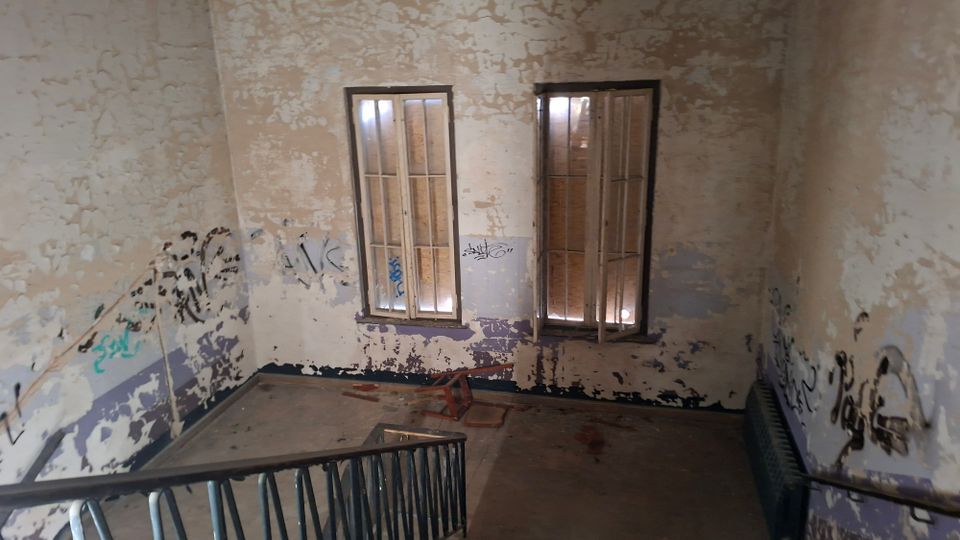 Innenraum eines stark verfallenen Treppenhauses mit abblätternder Farbe und Graffiti an den Wänden sowie einem Metallgeländer im Vordergrund. Zwei hohe Fenster mit Rahmen lassen Licht herein; der Boden ist karg und teilweise mit herumliegenden Teilen bedeckt.