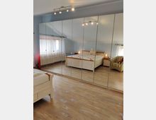 Schlafzimmer mit hellem Holzboden, einem großen Kleiderschrank mit Spiegeltüren, einem Holzbett und einem gepolsterten Sessel. Tageslicht fällt durch ein Fenster mit weißen Vorhängen.