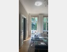 Helles Wohnzimmer mit hohen Decken, großen Sprossenfenstern und weißen Vorhängen, die einen Blick auf eine grüne Terrasse mit Sitzmöbeln freigeben. Der Raum ist mit einem weißen Sofa, schwarzen Stühlen und einem Glastisch auf einem Holzfußboden eingerichtet.