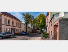 Straßenszene mit zweigeschossigen Wohnhäusern, teilweise mit Backsteinfassaden, geparkten Autos und Gehwegen, gesäumt von hohen Bäumen unter einem klaren blauen Himmel.