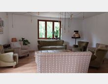 Wohnzimmer mit Retro-Einrichtung, bestehend aus einem grünen Sofa, mehreren gepolsterten Sesseln und einem gestreiften Sofa um einen rustikalen Holztisch. Im Hintergrund Fenster mit Blick ins Grüne, hängende Glühbirnen als Beleuchtung und Pflanzendekoration auf der Fensterbank.