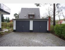 Ein gepflasterter Hof mit drei grauen Garagentoren an einem weißen Gebäude. Im Hintergrund ist ein Haus mit dunkelgrauem Satteldach sowie weitere Gebäude und Bäume zu sehen.