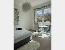 Helles Schlafzimmer mit modernem Arbeitsbereich, bestehend aus einem weißen Schreibtisch und einem Drehstuhl nahe großen Fenstern. Der Raum ist mit einer grauen Polsterbettbasis, hellen Dekorationen, darunter ein runder, federartiger Lampenschirm und Girlanden mit künstlichen Blumen, gestaltet.