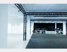 Ein heller Raum mit schwarzem Bodenbelag, ausgerüstet mit Metallträgern an der Decke und seitlich. Im Hintergrund befinden sich Arbeitsplätze mit Monitoren, Stühlen und Kamerastativen vor einer schwarzen Vorhangwand. Zwei Türen mit Beschriftungen "3D Body Scan" und "Voice Recording" sind an einer weißen Wand sichtbar.