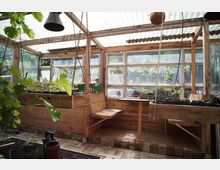 Innenraum eines rustikalen Gewächshauses mit Holzrahmen, großen Fenstern und einem Dach aus Wellplatten. Entlang der Wände stehen hölzerne Pflanzbeete mit Jungpflanzen; Seile und Lampen hängen von der Decke, der Boden ist teils mit Ziegeln ausgelegt.