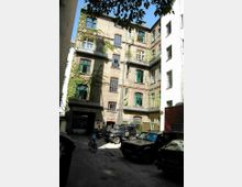 Backsteingebäude mit grünen Fensterrahmen in einem Innenhof, teilweise bewachsen mit Efeu. Im Vordergrund geparkte Autos und ein Fahrradfahrer, links ein Torbogen, der den Innenhof mit einer anderen Straße verbindet.