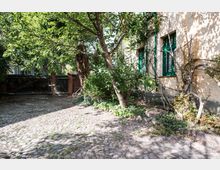 Ein gepflasterter Innenhof mit einem Baum in der Mitte und dichter Vegetation am Rand. Rechts befindet sich eine gelb-weiße Hausfassade mit grünen Fensterrahmen, im Hintergrund ist ein Holztor mit Ziegelpfosten zu sehen.