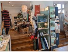 Ein kleiner Laden mit verschiedenen Geschenkartikeln, Dekorationen und Kleidung. Zu sehen sind Holzregale mit Kissen, Karten und Keramik, ein Kleiderständer mit bunten Kleidungsstücken und eine Holztreppe, die zu weiteren Verkaufsbereichen führt.