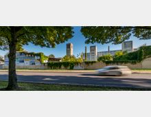 Straßenansicht eines großen, historischen Fabrikgebäudes mit einer zentralen Turmuhr und umgebendem Bewuchs auf einer Mauer. Im Vordergrund verläuft eine Straße, entlang derer ein Auto vorbeifährt, flankiert von Bäumen, die Schatten spenden.