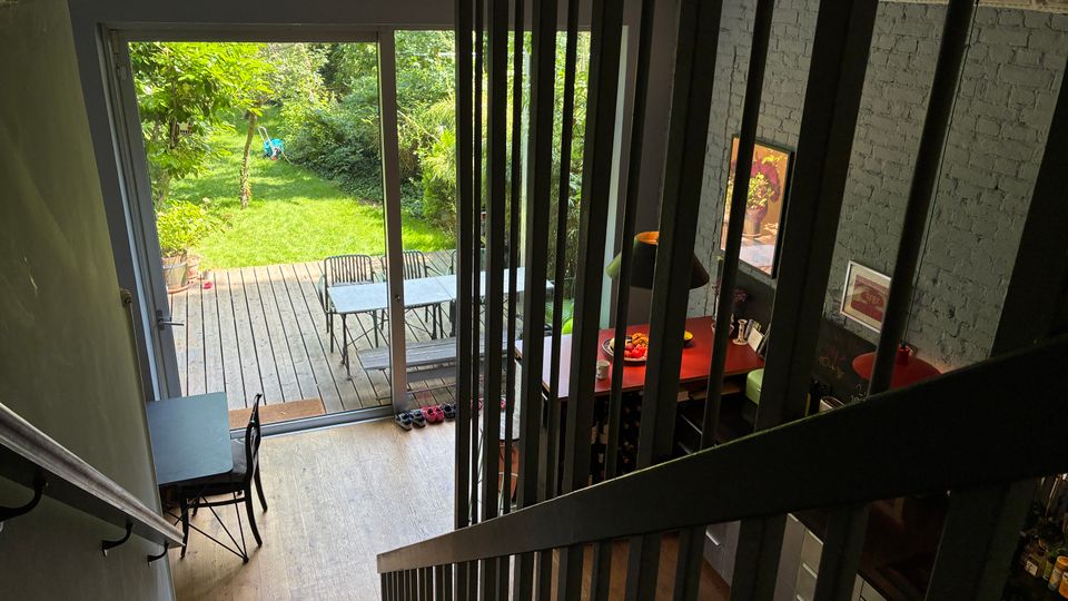 Blick von einem Treppenaufgang in einen offenen Wohnbereich mit vertikalen schwarzen Geländerstreben und einer Wand in Backsteinoptik. Große Glasschiebetüren führen auf eine Holzterrasse mit Tisch und Stühlen und weiter in einen grünen Garten mit Bäumen und Sträuchern.