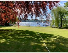 Grüne Wiese mit schattenspendenden Bäumen und roten Blättern im Vordergrund, Blick auf ein funkelndes Gewässer mit Booten und einer dichten Waldkulisse im Hintergrund. Am Ufer stehen Sitzbänke, und ein Wasserschlauch verläuft über den Rasen.