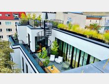 Modernes Stadthaus mit begrüntem Flachdach, einer Wendeltreppe, die vom Innenhof zur Dachterrasse führt, und großen bodentiefen Fenstern. Der Innenhof ist mit Pflanzen, Sitzmöbeln und einem Holztisch gestaltet, umgeben von benachbarten Wohnhäusern.